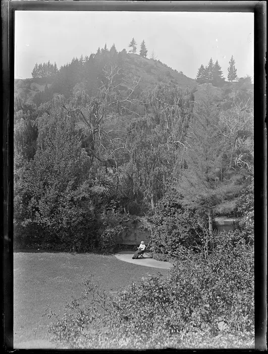 Lydia Williams, sitting and reading a book in a garden, including surrounding bush and trees, [Nelson]
