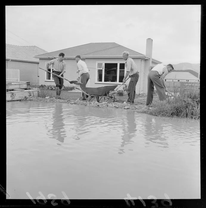 Water main bursts at Wainuiomata with unidentified men shovelling dirt to create a barrier around a house, Lower Hutt, Wellington Region