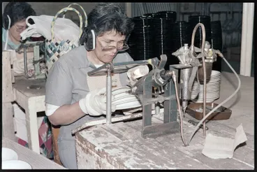 Image: Negative - Worker sitting at mounted machine, holding stack of plates applying colour or similar