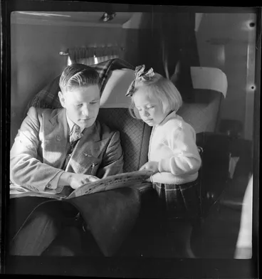 Image: Young child Ms Shandler, a passenger on National Airways Corporation Douglas DC-3 flight from Auckland to Fiji, reading a newspaper with an unidentified male passenger