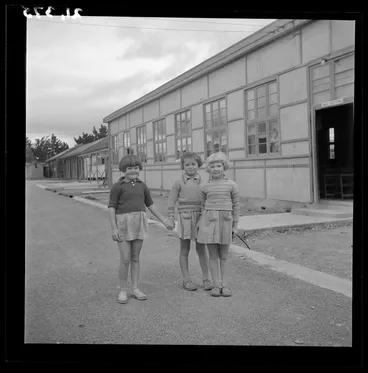 Image: Three young girls pose outside the auditorium (sala odczytowa) at a Polish refugee camp, Pahiatua