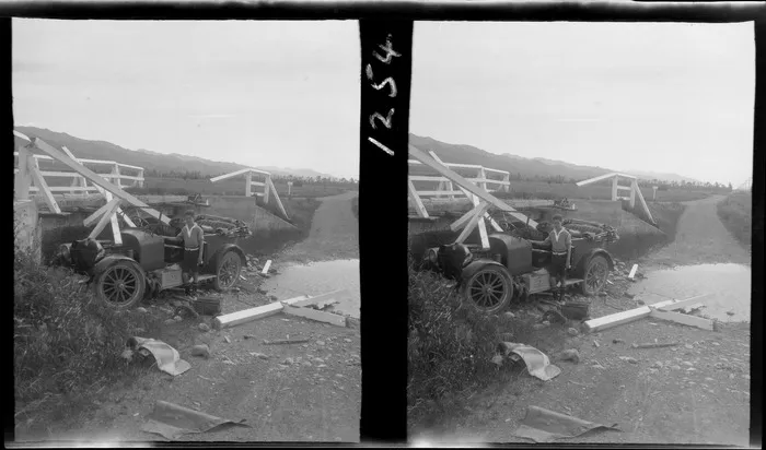 Unidentified teenaged boy beside Ford touring car that has crashed through the barriers of a small wooden bridge on an unknown country road