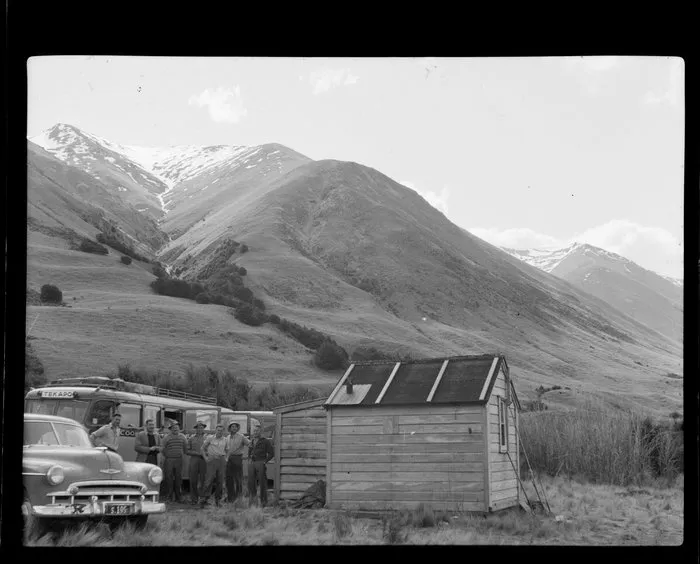 Lake Ohau, Central Otago, showing a group of men with Vauxhall Velox car and Tekapo bus, beside a drovers cabin on tussock ground, with snow covered mountains and creek beyond