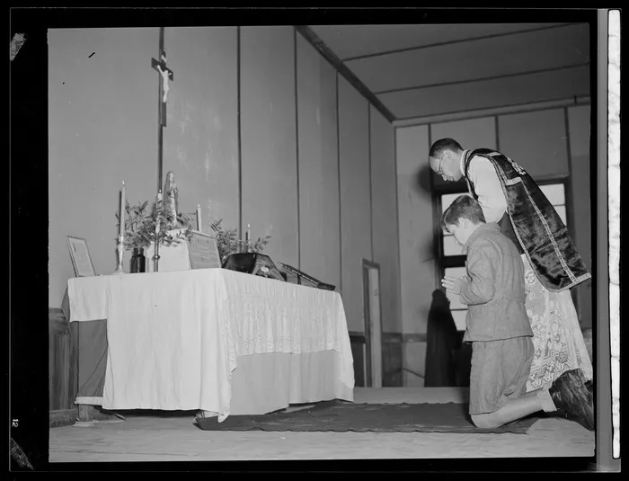 Father M Wilniewczyc and an unidentified boy praying at an altar in auditorium (sala odczytowa), at Polish children's refugee camp, Pahiatua