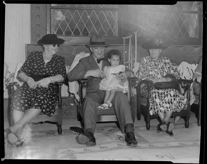 Sir Peter Buck sitting next to two women in the meeting house and holding a little girl, Ngaruawahia, Waikato