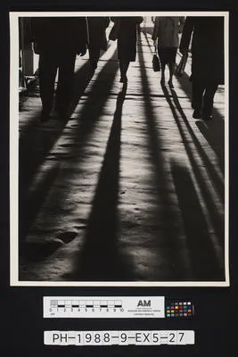 [Shadows cast by pedestrians walking along a paved footpath]