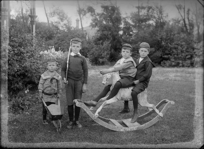 Outdoors on grass with trees beyond, an unidentified family portrait with young brothers with caps, small wooden wheelbarrow, toy rifle and rocking horse, older boy with Eton shirt collar and bow tie, others in large collar shirts, probably Christchurch region