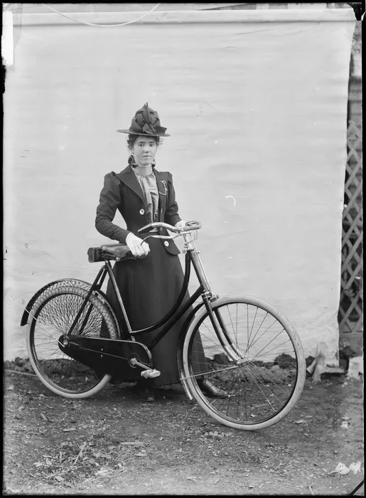Unidentified young woman, with a bicycle, outdoors, with a backdrop, probably Christchurch district