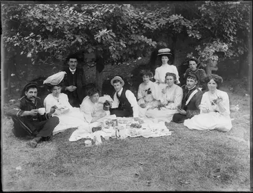 Image: Group picnicking, showing unidentified men, some in military uniforms, and women in tea dresses, sitting around a cloth laid with food, possibly Christchurch district