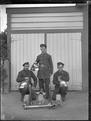 Image: Three firemen in front of the Petone Fire-Brigade Station, with helmets, hoses and other pieces of equipment