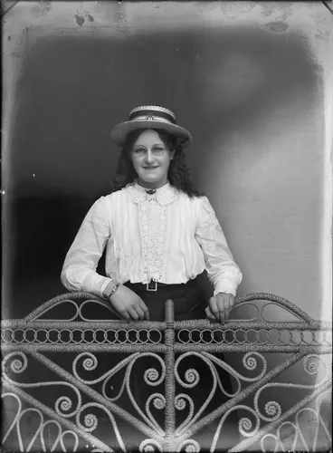 Image: Studio portrait of unidentified young woman wearing spectacles and a straw boater hat standing next to a cane chair, probably Christchurch district
