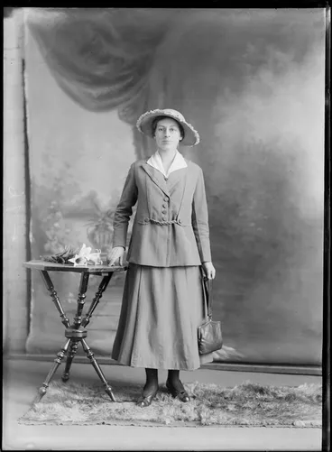 Image: Studio unidentified portrait of a young woman with glasses in a large collar blouse, matching jacket and skirt and hand bag, standing next to a table with a flower, Christchurch