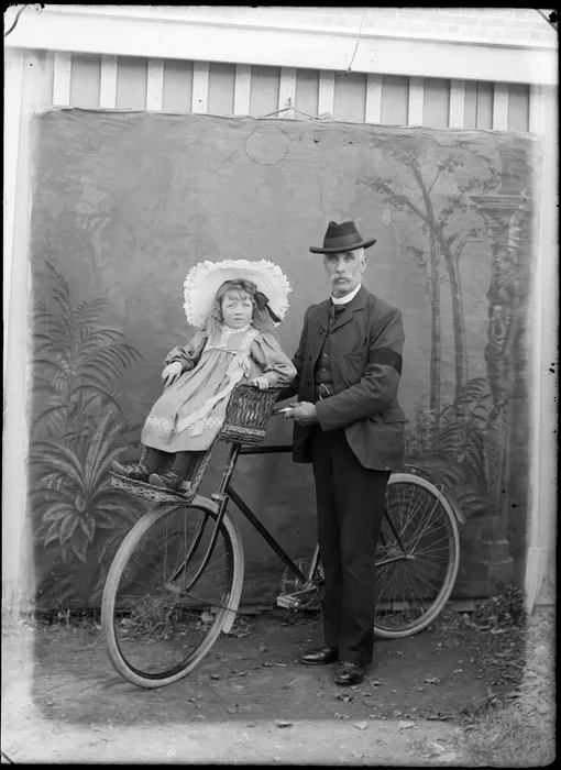 Outdoors portrait of man holding a bicycle and a young girl with large hat is on a front handlebars wicker child carrier, probably Christchurch region