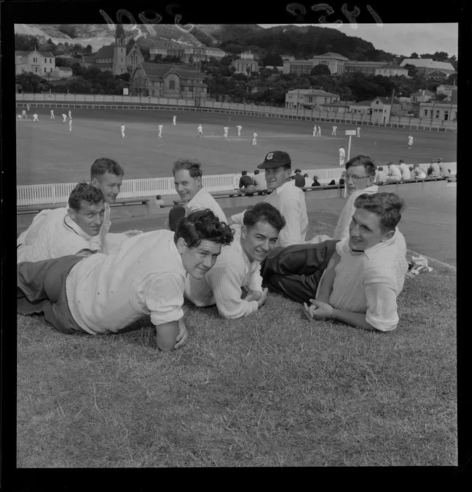 Unidentified cricket players on embankment, with game of cricket being played in background, Basin Reserve, Wellington