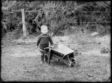 Image: Outdoors in backyard area with wire fence and gorse behind, unidentified small boy with small wooden wheelbarrow, probably Christchurch region