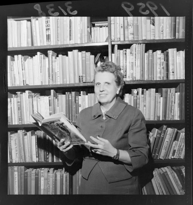 Miss A Lohrer, with book in hand, standing next to book case