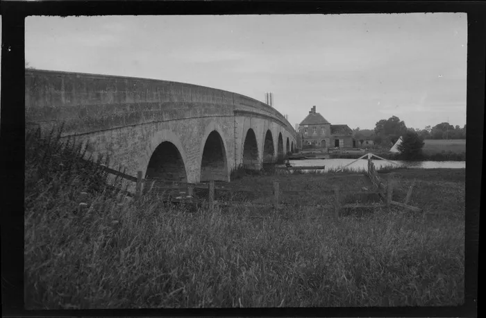 An eight arched stone bridge over Gave de Pau river, a man fishing in a boat, a stone house and farmland beyond, Pau, South-west France