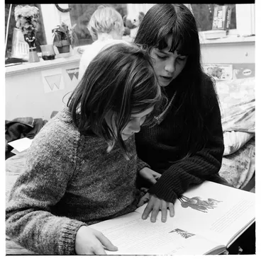 Image: Two young girls reading a book together, and playing with toys