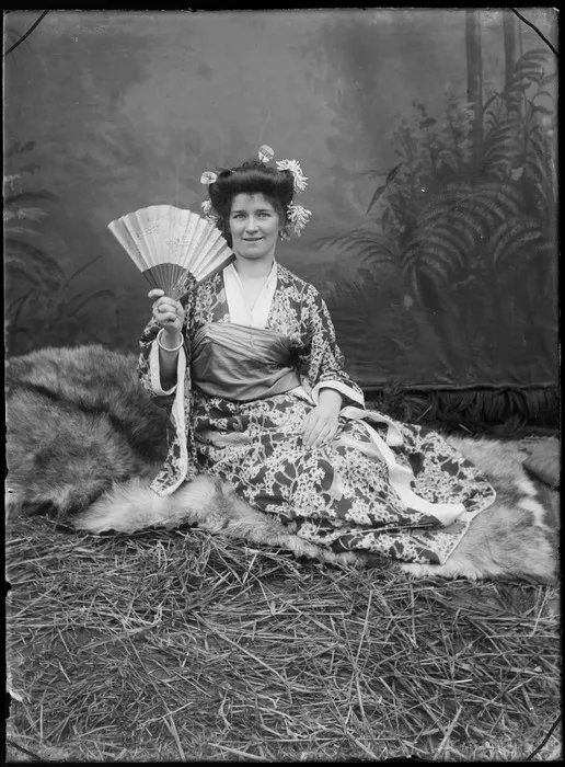 Studio portrait of an unidentified young woman dressed in Japanese silk kimono, with fan in hand and flowers in her hair, sitting on fur rug and straw, Christchurch