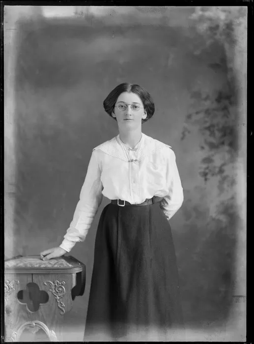 Studio unidentified portrait of a young woman with glasses, in a large lace collar cotton shirt with jewelled bar and silver fern brooches, standing beside a wooden highchair, Christchurch