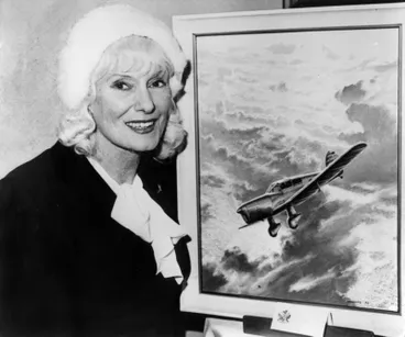 Image: Jean Batten standing in front of the cover illustration produced by Lynn Williams for the republication of Jean Batten's biography