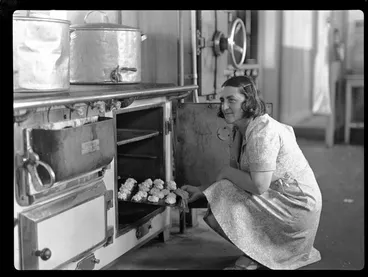 Image: Portrait of an unidentified female TEAL Catering Staff member putting a tray of scones into an oven within a kitchen, Mechanics Bay, Auckland City