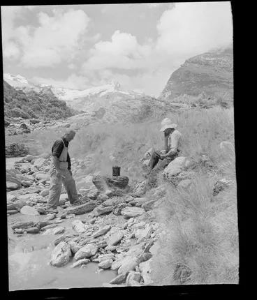 Image: Two unidentified men lighting a campfire next to a river, Fox Glacier, West Coast Region