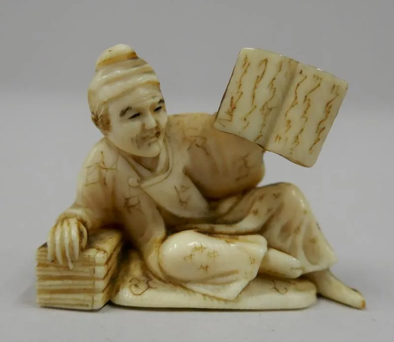 Netsuke: Kajin [A Poet]; Netsuke in the form of a man reclining on a pile of books, holding up another.