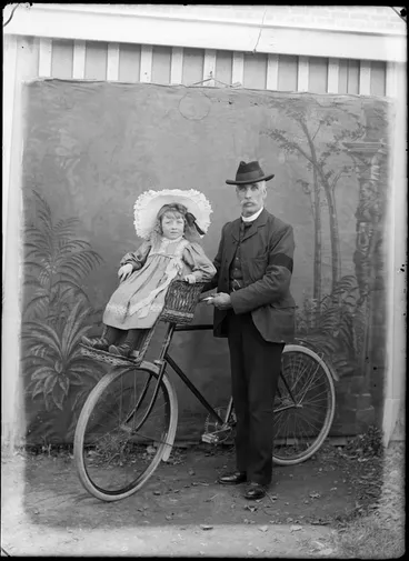 Image: Outdoors portrait of man holding a bicycle and a young girl with large hat is on a front handlebars wicker child carrier, probably Christchurch region