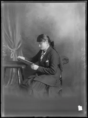 Image: Studio portrait of an unidentified girl wearing a school uniform, sitting in a chair, reading a book, possibly Christchurch district