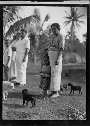 Image: A portrait of a local Tongan mother and child with young dogs, with family members or neighbours behind, Tonga