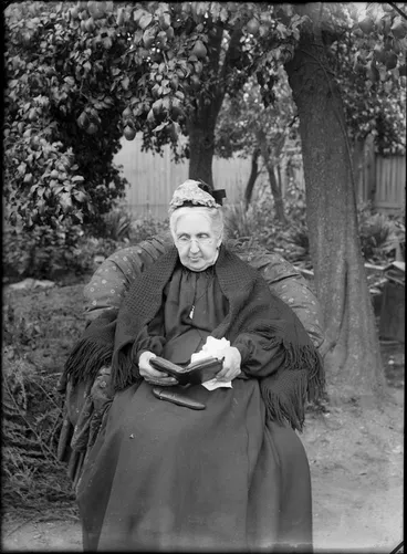Image: Outdoors backyard area, an unidentified elderly woman sitting under a pear tree with glasses, shawl and lace cap reading a book, with glasses case in her lap, probably Christchurch region