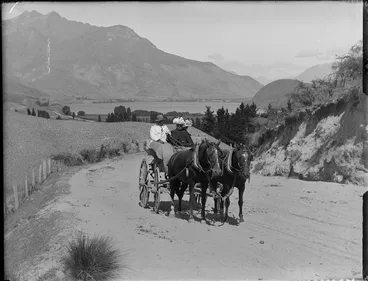 Image: Lydia Williams and an unidentified couple on a two horse drawn wagon on a dirt hill road through farmland with Lake Wakatipu behind, Queenstown District, Central Otago Region