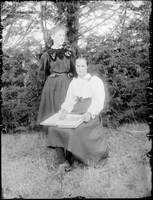 Group portrait of two unidentified young women, one sitting with a book on lap, on grass slope with tall trees behind, probably Christchurch region