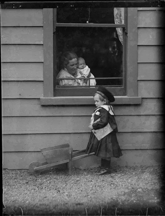 Lydia Williams holding Owen sitting by a window with Edgar outside playing with a toy wheelbarrow, at their home 'View Bank', Maitland Street, Dunedin