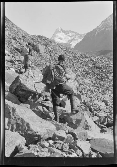 Edgar Williams and an unidentified climbing companion climbing a moraine slope, mountains rising above, near Lyell Glacier, Southern Alps, Canterbury Region