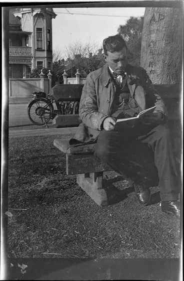 Image: An unidentified man sitting on a wooden bench beneath a tree, reading and smoking a pipe, motorbike parked in street behind, large wooden building opposite, probably Christchurch
