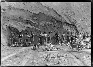 Image: Road construction workers stop for a break while loading wheelbarrows with rocks and dirt, shows most men holding spades