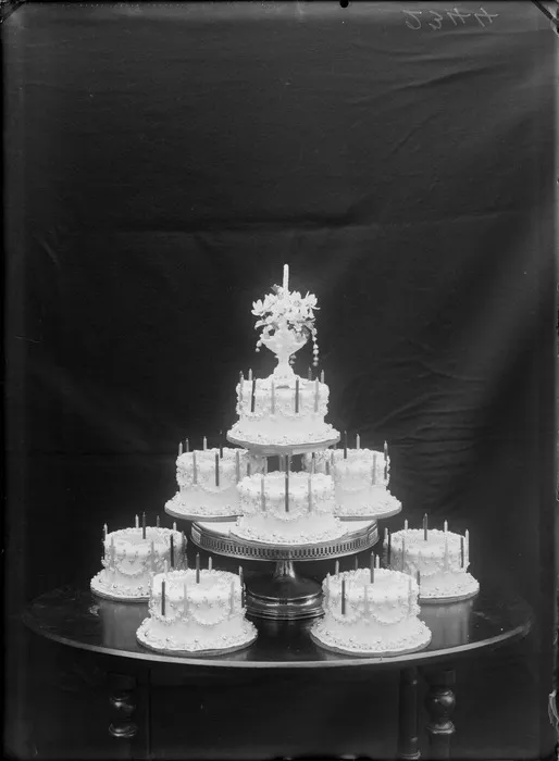 Eight identical birthday cakes with candles arranged on a table, top most cake with vase holding flowers, probably Christchurch region