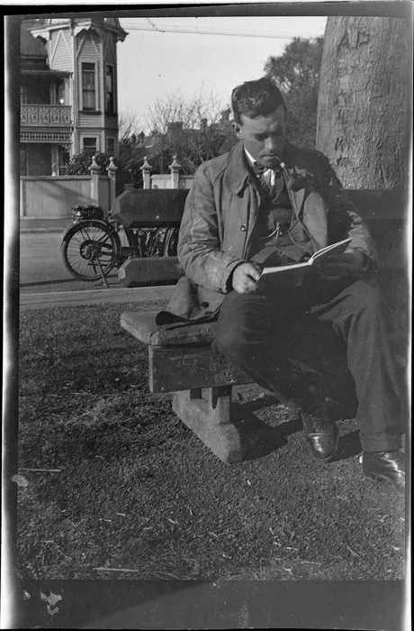 An unidentified man sitting on a wooden bench beneath a tree, reading and smoking a pipe, motorbike parked in street behind, large wooden building opposite, probably Christchurch