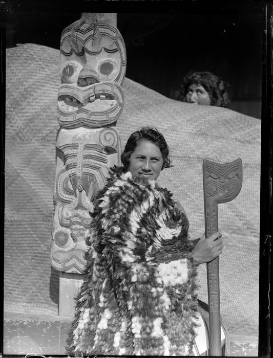 Tarihira Mihinui dressed in a feather cloak holding a tewhatewha, sitting by a carved wooden pou, Otūkou marae, Lake Rotoaira