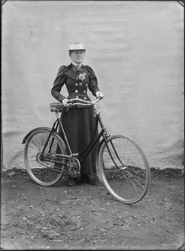 Image: Outdoors portrait with false backdrop, an unidentified woman in dark double breasted jacket with large white buttons and flowers, tie and straw hat, standing holding women's bicycle, probably Christchurch region