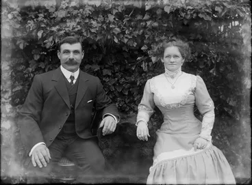 Image: Outdoors couples portrait in front of ivy hedge, husband with stiff arundel shirt collar and handlebar moustache, sitting with wife in embroidered dress, with necklace, brooch and glasses, probably Christchurch region