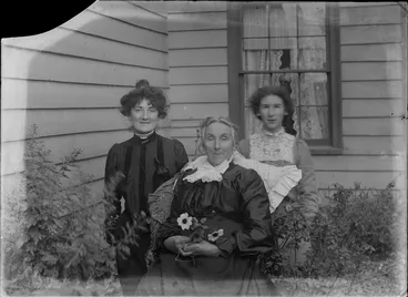 Image: Three unidentified women, including a mature woman sitting in a wicker chair holding a posy and spectacles, in a garden, weatherboards and window of a house behind, possibly Christchurch district