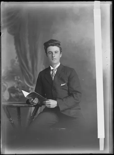 Image: Studio portrait of unidentified man sitting, leaning on a high table holding a book, probably Christchurch district
