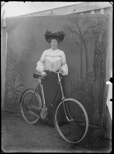 Image: Outdoors portrait in front of false backdrop, unidentified woman with lace blouse, necklace and large hat with ribbons, standing with women's bicycle, probably Christchurch region