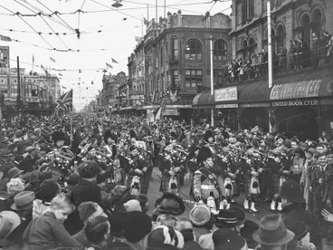Image: Central City. Celebration. Victory in Europe or VE Day. Led by the Skirl of the Pipes Men March Through the "Bottleneck" and on to Cathedral Square. Crowds of Onlookers. Note This was celebrated on the 9 May, Two Days After the Actual Surrender As the Deputy Prime Minister, W. Nash, waited to Hear it Officially From Churchill. Colombo Street, Christchurch, Canterbury, New Zealand.