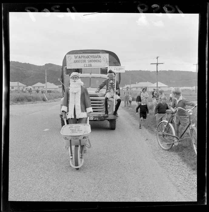 Parade at Wainuiomata, Wellington, for swimming baths fund, showing unidentified man dressed in a Santa Claus costume with a wheelbarrow, in front of a bus which reads 'Wainuiomata Amateur Swimming Club'