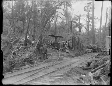 Image: [William Williams?] standing on railway lines at a logging camp with a winch cable steam engine and carriages under trees, Kakahi District, Manawatu-Whanganui Region