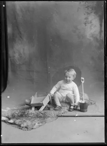 Image: Studio portrait of an unidentified small boy, sitting on a fur rug on floor with a wooden toy wheelbarrow and a spade and pail, possibly Christchurch district